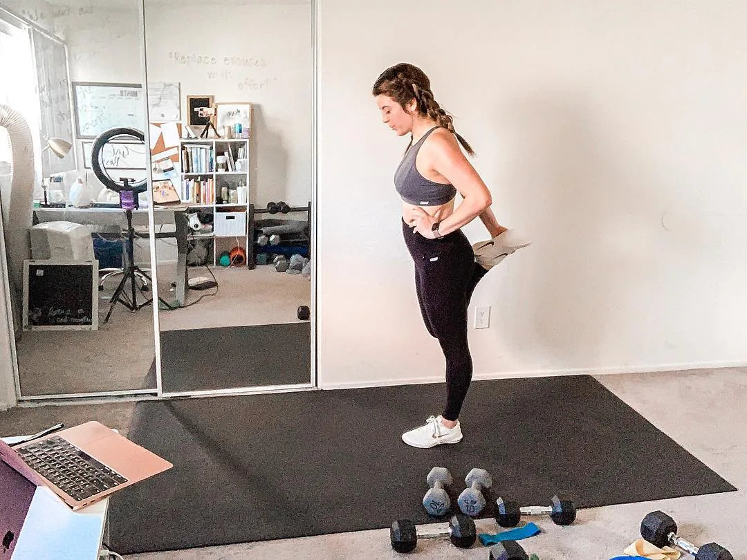 Woman stretching in sneakers on a large black mat in her home office, dumbbells on the mat