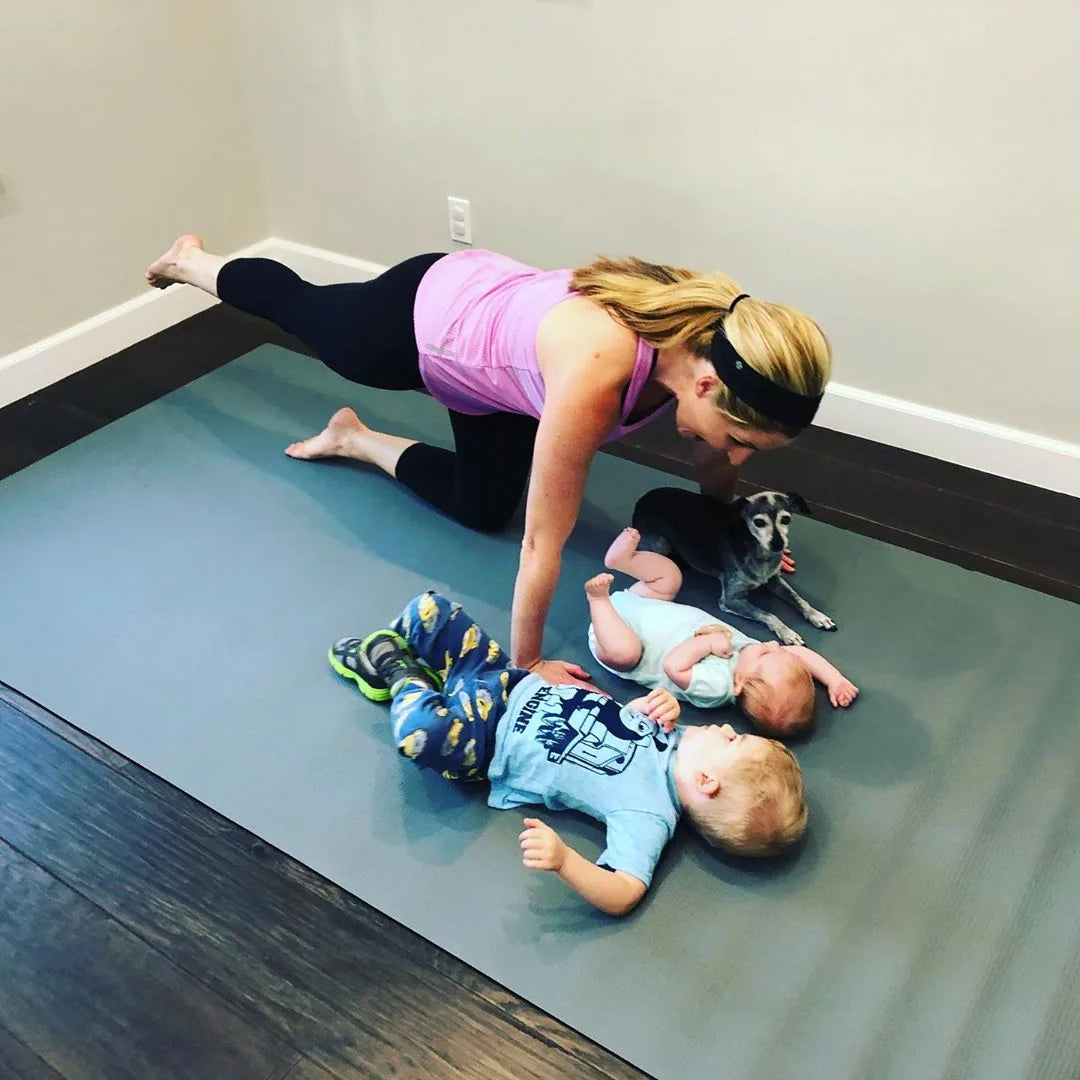 Mom in a plank over her two babies on a large grey mat on a dark wood floor, small dog beside them