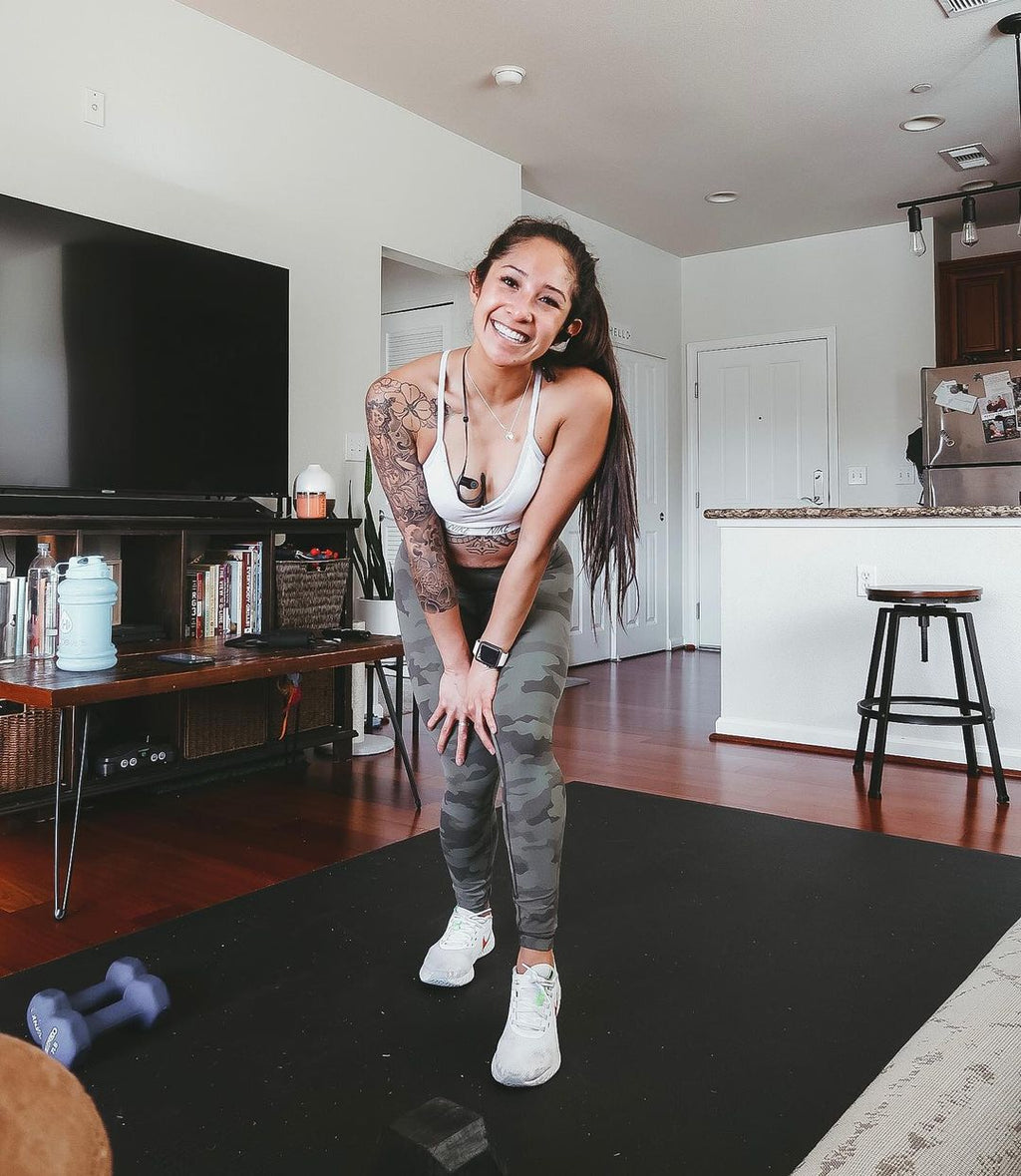 Smiling woman in sneakers on a large black exercise mat in an open-plan living room, dumbbells beside her