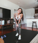 Smiling woman in sneakers on a large black exercise mat in an open-plan living room, dumbbells beside her
