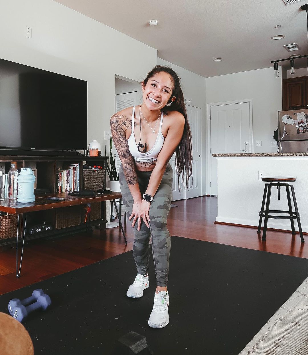 Smiling woman in sneakers on a large black exercise mat in an open-plan living room, dumbbells beside her