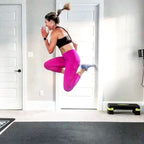 Woman in sneakers jumping high above a large black exercise mat at home