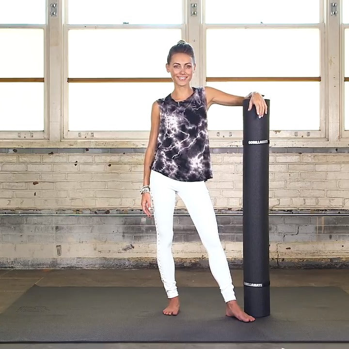 Smiling woman standing on a large black yoga mat with a rolled mat beside her