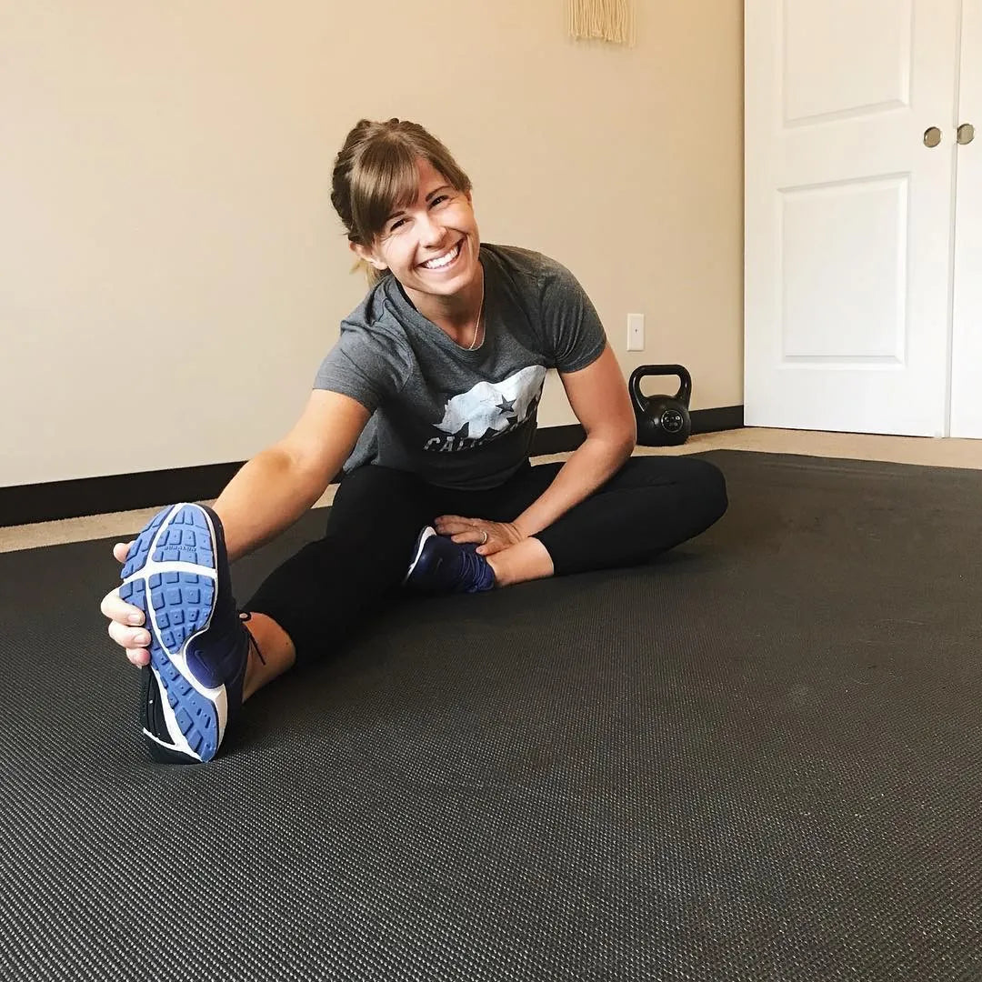 Smiling woman in sneakers stretching on a large black exercise mat at home, a kettlebell behind her