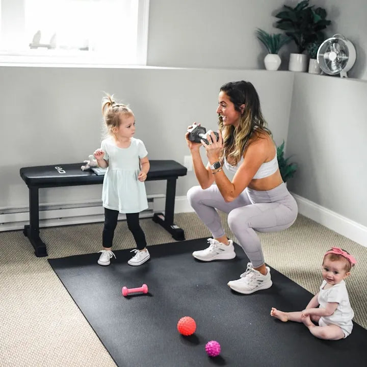 Mom squatting on a black mat while her toddler and baby play beside her
