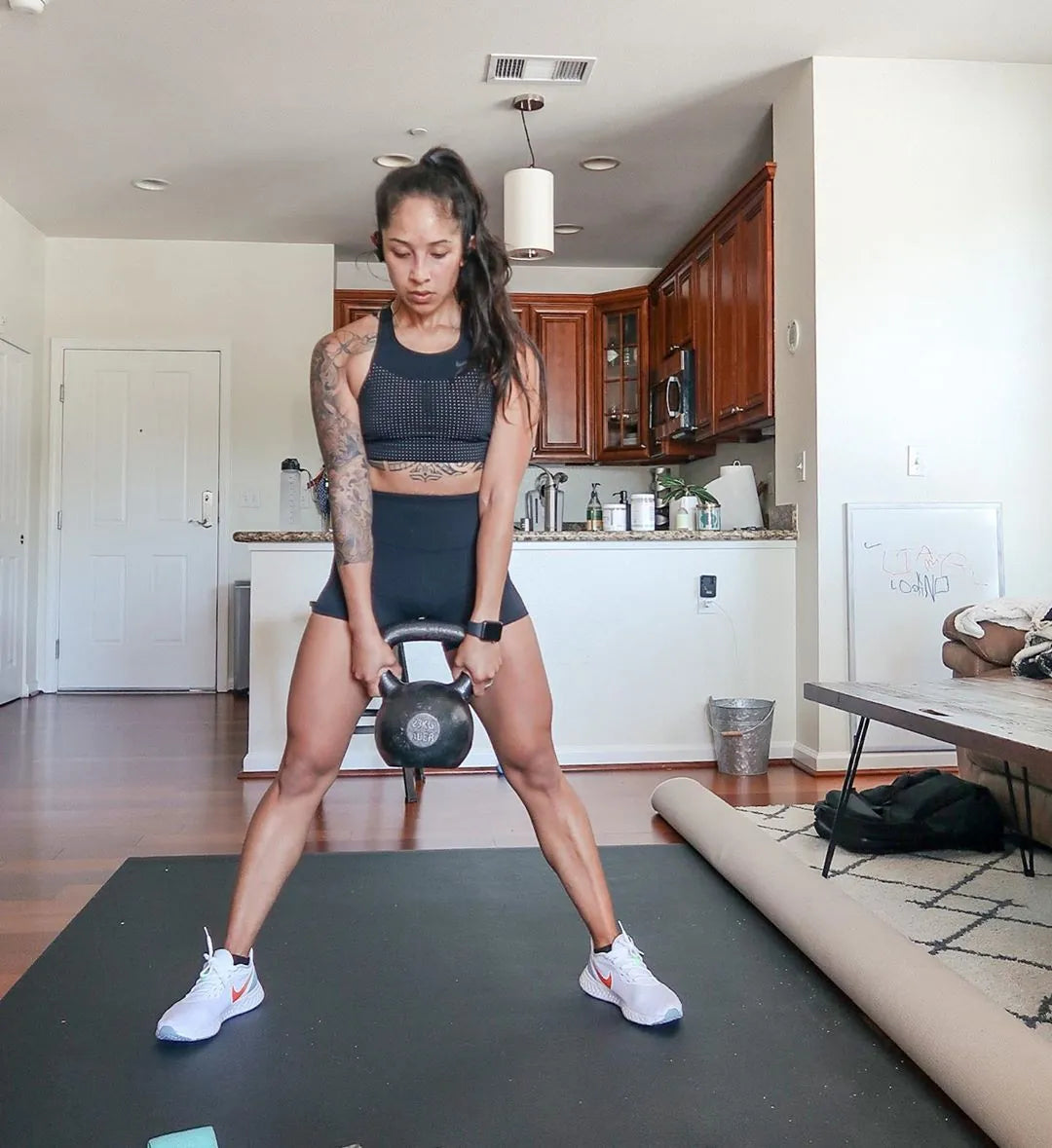 Woman in sneakers lifting a kettlebell on a large black exercise mat in an open-plan living room