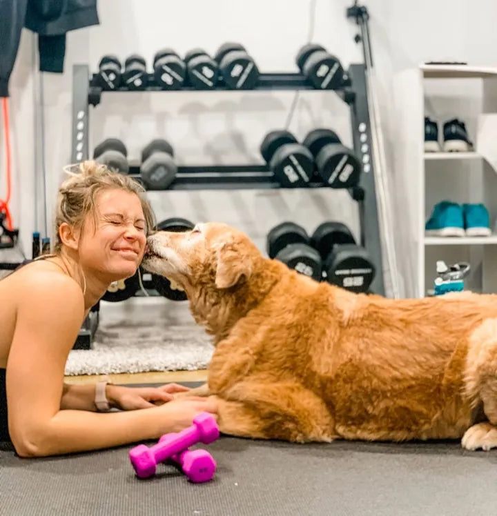 Woman lying on a black mat nose to nose with a golden retriever, dumbbell rack behind