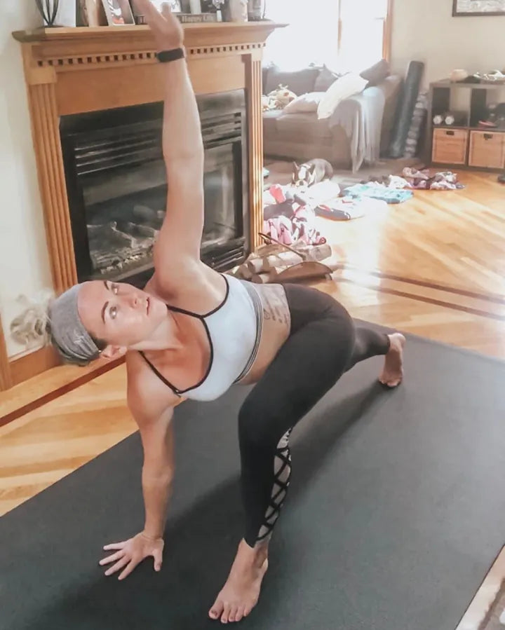 Barefoot woman in a twisting side lunge on a large black mat by her fireplace